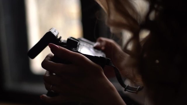 Close-up of young female photographer's hands holding camera and looking at photos in professional dark studio, against the window. Concept of creative work in photo studio. Shooting in slow motion.