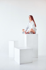 Pensive girl sitting on the white cube, white background.