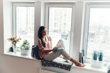 Attractive young woman in cozy pajamas reading a book while resting on the window sill at home