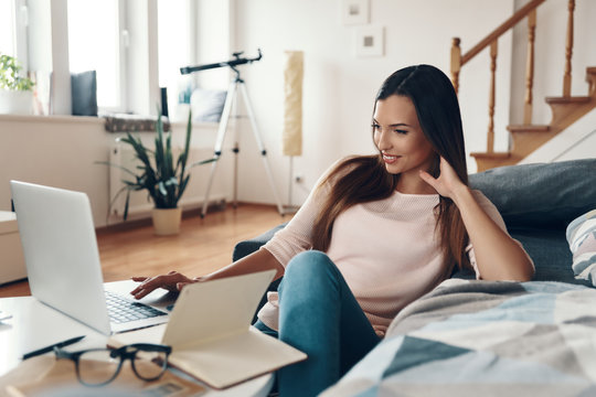 Cute young woman in casual clothing using laptop and smiling while resting at home
