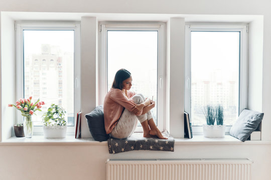 Attractive young woman in cozy pajamas thinking about something while resting on the window sill at home
