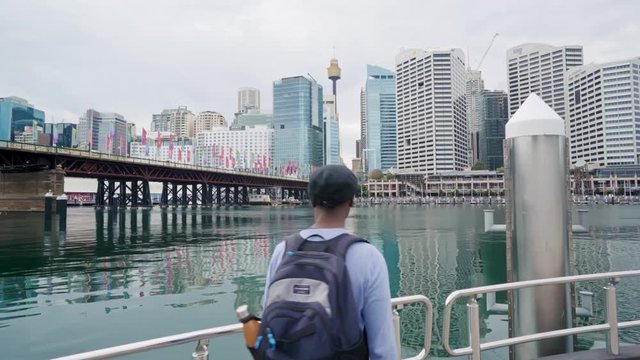 Over the shoulder shot of black tourist walking up to the edge of the Darling Harbour pier before turning around back towards the camera in surprise and awe. 
