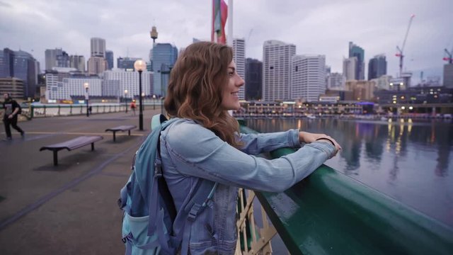Female backpacker smiles at the camera before admiring the view of Darling Harbour and the Sydney cityscape from Pyrmont Bridge
