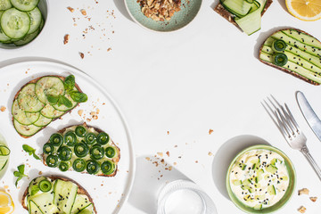 top view of fresh cucumber toasts on plate near lemon, water, cutlery and yogurt on white background