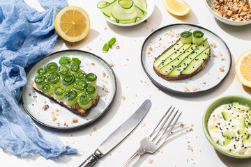 fresh cucumber toasts on plates near cutlery, yogurt and lemon on white background