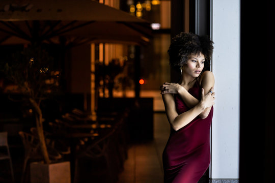 Young African American black woman at night in street. Woman posing near the window of a street cafe, dressed in a cute red dress. Nightlife, fashion and style concept