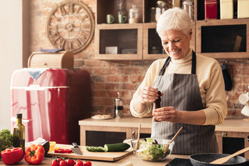 Happy elderly woman seasoning fresh salad with pepper mill © Prostock-studio