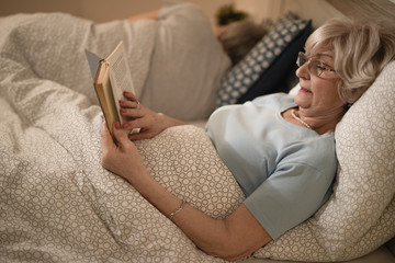 Mature woman lying down in bed and reading book