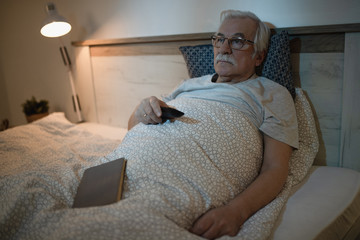 Pensive mature man watching TV while resting in bed at night.