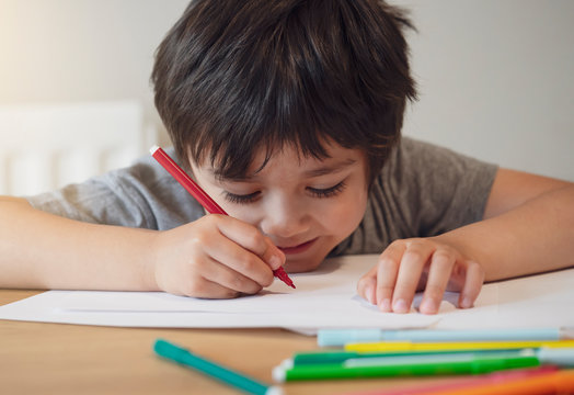 Selective Focus Of School Kid Boy Siting On Table Doing Homework, Happy Child Holding Red Pen Writing Or Drawing On White Paper, Elementary School And Home Schooling, Education Concept