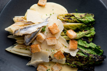 Caesar Salad. Grilled Romaine lettuce leaves topped with classic Caesar salad dressing and garnished with butter garlic croutons shaved parmesan cheese and whole anchovies. Classic American Salad.