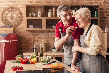 Loving aged man feeding his wife with cucumber in kitchen