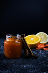 Orange carrot jam in a small jar on a black background with ingredients in the background, oranges and carrots