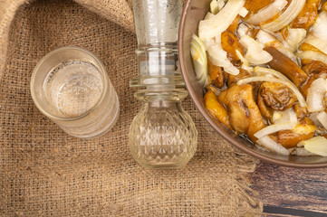 Salted mushrooms with onions in a ceramic bowl, a glass of vodka and a decanter on a background of rough homespun fabric. Homemade preparations, Rustic treats. Close up.