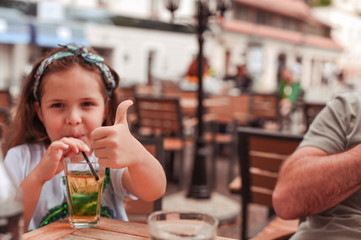 Little girl in a cafe on the street drinks juice. child has fun, shows hands gestures
