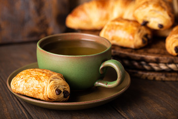 Green cup of tea with mini chocolate bun, puff pastry on old wooden table. Tasty tea break concept, copy space.