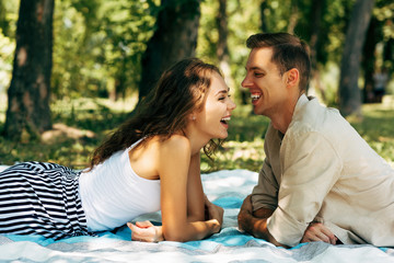 Side view image of happy couple in love laughing and dating outdoors at the park on a sunny day. Young man ansd woman lying on the blanket on the grass during picnic in the nature. Happy relationship