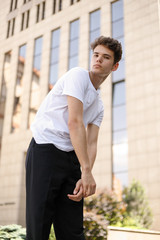 Elegant young man in a fashionable black shirt in a white stylish t-shirt in black pants with a trendy hairstyle rests near a modern business center. Attractive guy in the street on a summer day.