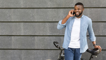 Joyful black guy standing with bike and talking on smartphone outdoors