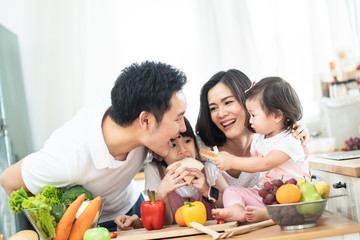 Lovely cute Asian family making food in kitchen at home. Portrait of smiling mother, dad and children standing at cooking counter. kid feeding dad some bread with smile. Happy family activity together