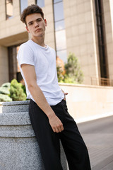 Elegant young man in a fashionable black shirt in a white stylish t-shirt in black pants with a trendy hairstyle rests near a modern business center. Attractive guy in the street on a summer day.