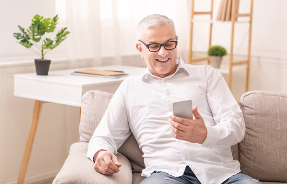 Cheerful aged man using smartphone sitting on sofa