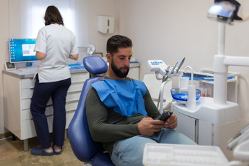 Fototapeta premium Male patient being looking at the smartphone while the female dentist prepares to work.