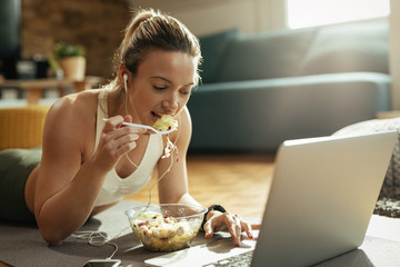 Female athlete eating salad and using laptop while relaxing at home.