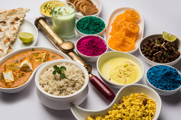 Happy Holy concept  showing Indian assorted lunch food like chicken, paneer butter masala, naan, jeera rice, black chana fry, jalebi, fujiyama, thandai and Farsan with holi colours and pichkari 
