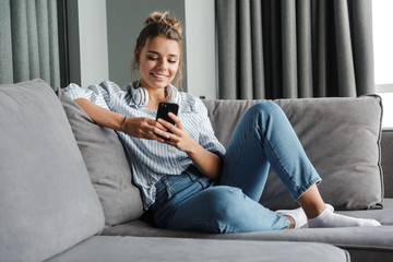 Image of smiling nice woman using cellphone while sitting on sofa
