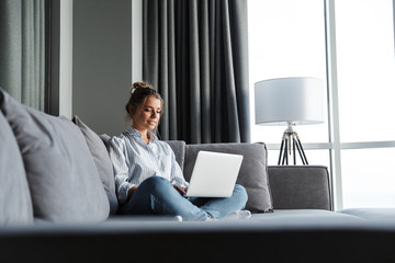 Image of pleased woman smiling and using laptop while sitting on couch