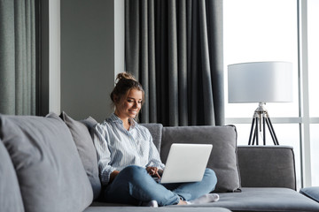 Image of joyful woman smiling and using laptop while sitting on couch