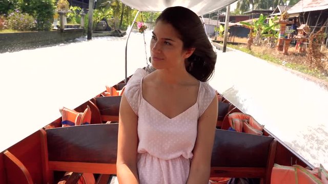 Beautiful tourist woman sightseeing from a boat in the floating market Damnoen Saduak in Ratchaburi, Thailand. Slow motion of her long hair moved by the wind. Holidays and travel in Asia concept.