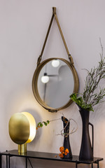 Round mirror is hanging on a leather rope belt, a dressing table with a gold lamp and vases with flowers
