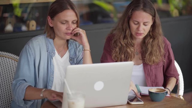 Happy women sitting at table in coffee shop and work together using laptop