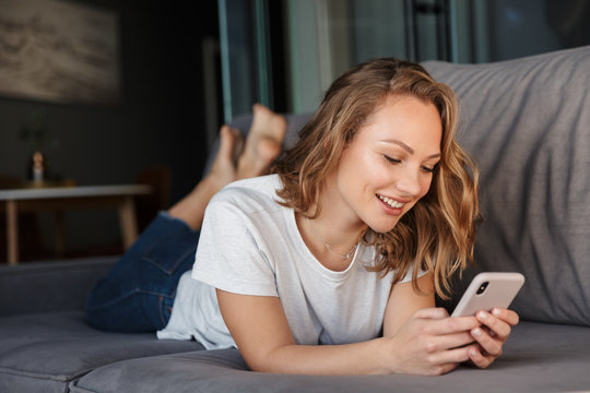 Image of happy woman smiling and using cellphone while lying on sofa