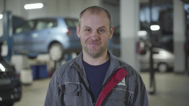 Close-up of adult Caucasian maintenance engineer taking off cap and wiping forehead with hand. Smiling male auto mechanic posing in car repair shop. Man working at service station.