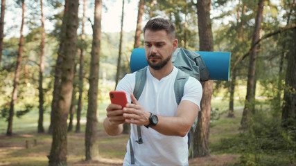 Handsome man with beard and backpack in forest uses speak on phone camera show around gps application orientation tourist travel hike lost sun communication telephone slow motion