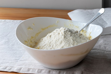 A white bowl with cottage cheese dough, on which a hill of flour is poured, stands on a linen tablecloth on a wooden table. The process of making curd bagels or croissants