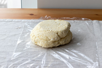 Fototapeta na wymiar Raw curd dough with bits of cottage cheese on a plastic wrap on a linen tablecloth on a wooden table. The process of making curd bagels or croissants