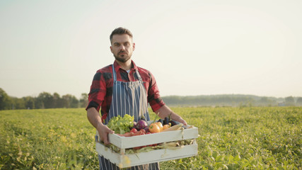 Close up handsome farmer is holding a box of organic vegetables walk look at camera at sunlight...