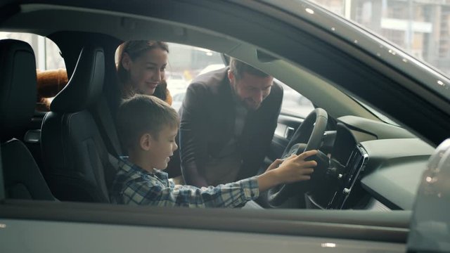 Joyful kid is sitting in new car holding steering wheel having fun while parents are buying automobile in dealership. People and consumerism concept.