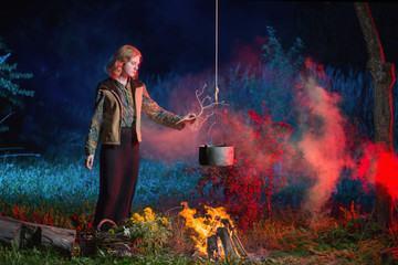 young witch by  fire in night forest prepares  magic potion