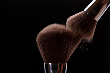 Make-up brush with beige powder explosion on black background. Creative splash. Professional powder. Cosmetics brush releasing a cloud of sparkling face powder. Copy space. Beauty and makeup concept