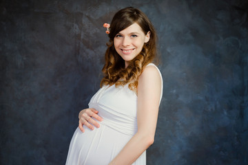 Fototapeta na wymiar Beautiful elegant pregnant woman in white dress smiling standing over dark backdrop. Maternity, happy pregnancy concept.
