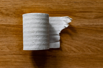 A roll of clean, plain toilet paper, cheap quality, dirty color, lies on a brown wooden table, slightly unwound and with the end torn off