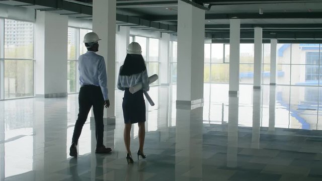 Rear view shot of two designers with drafts and helmets going through hall of new building and inspecting constructions