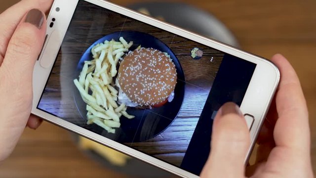 A Girl's Hands Take Photos with a Cell Phone of a Fast Food Burger With French Fries on a Wooden Surface