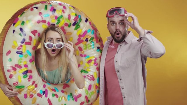 Excited fun young couple posing with inflatable float donut on orange background. Portrait of curious man and woman staring at camera surprised eyes wide enjoying summer vacation.