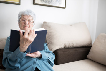 Serious senior white-haired Caucasian woman relaxing in her apartment and reading a book. Loneliness and solitude concept